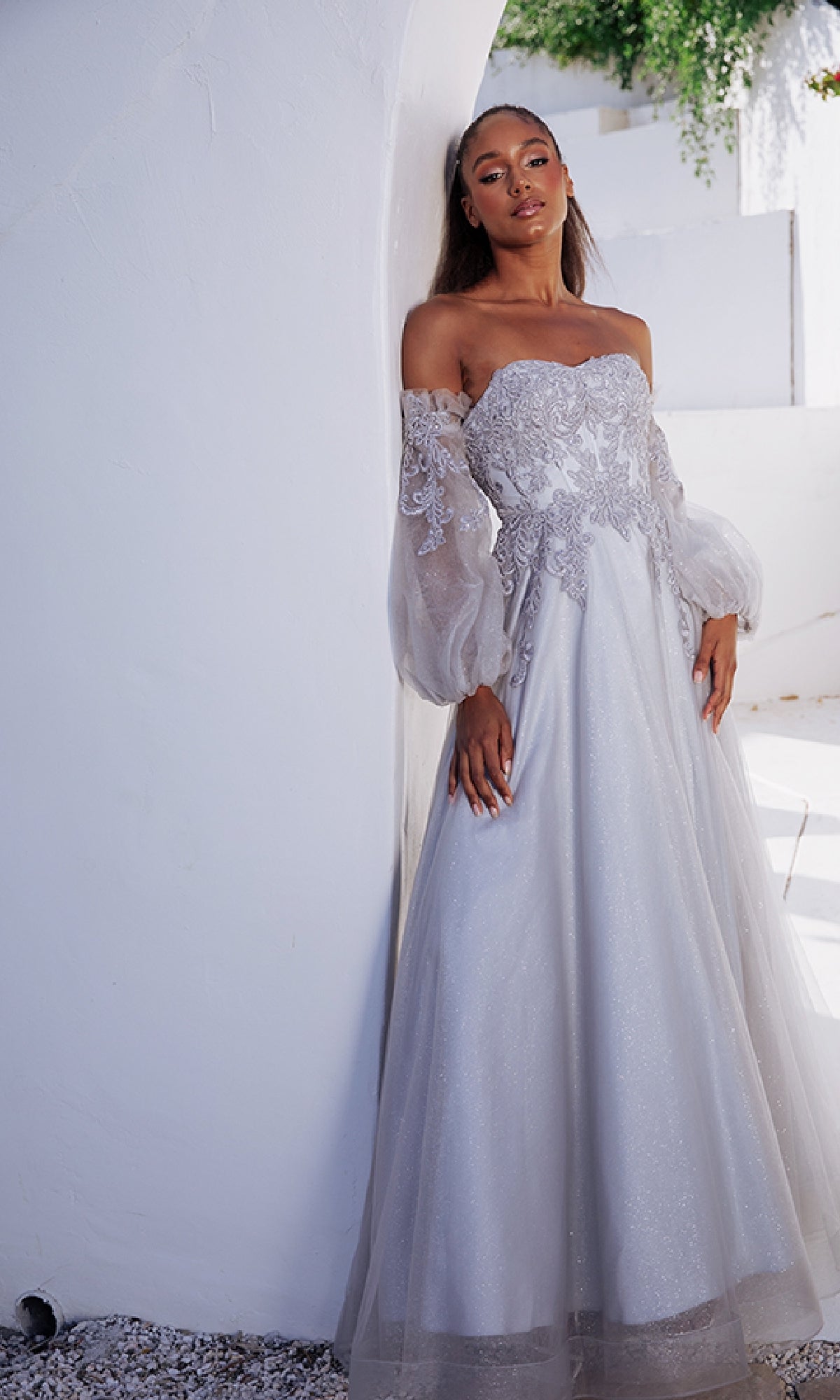A woman in the Eureka 9505 Long Sleeve Strapless Long Prom Gown, featuring an embroidered sweetheart bodice and detachable balloon sleeves, poses by a curved white wall in a sunlit outdoor setting.